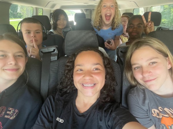 Group of 4-H'ers in vehicle 3 girls in first row bow with finger to lips in second row girl grinning and boy making peace sign.